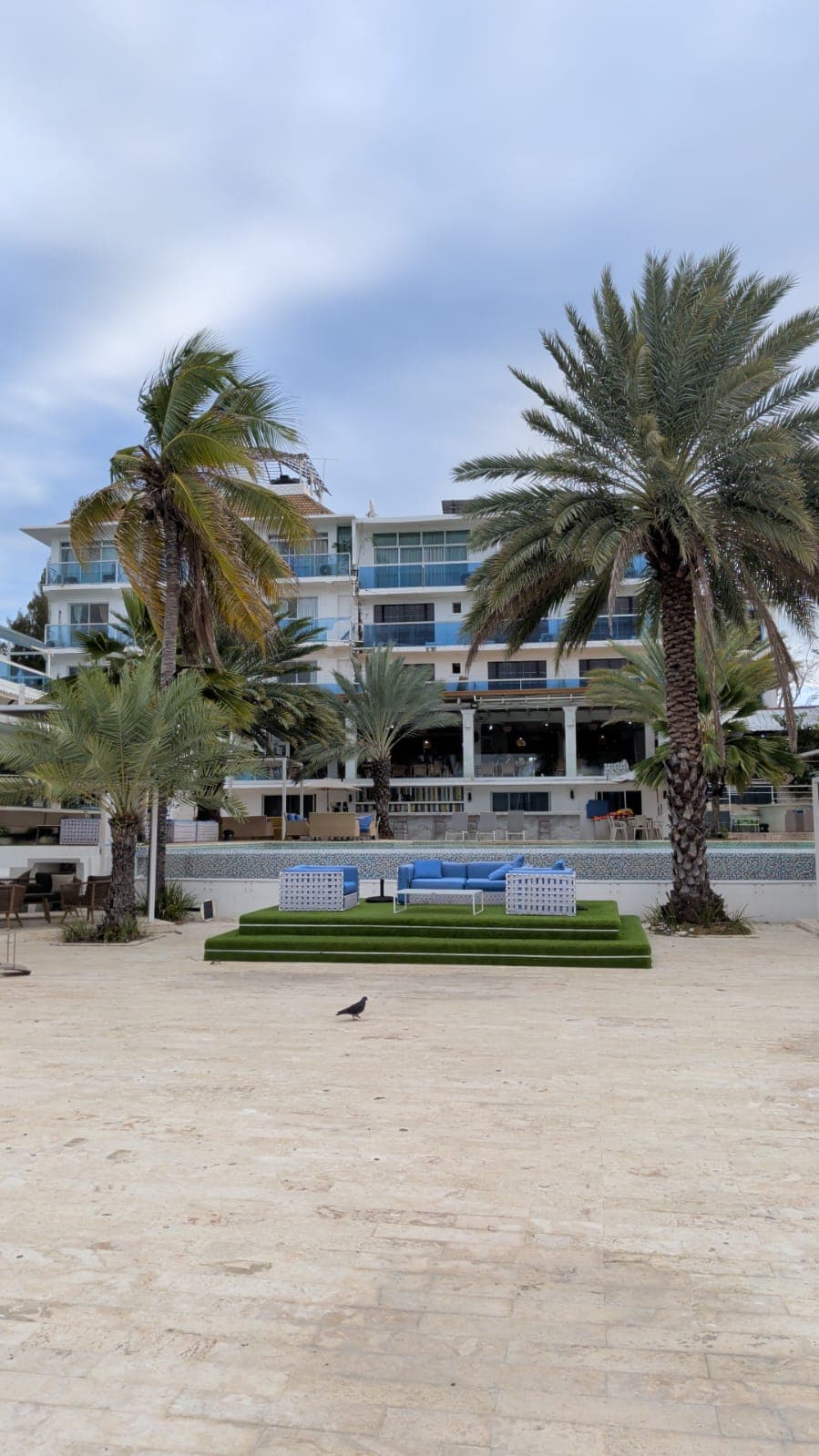 Hôtel blanc moderne avec balcons bleus, palmiers et canapés bleus sur une plateforme verte.